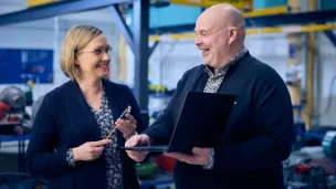 Kaksi hymyilevää henkilöä keskustelemassa teollisuusympäristössä, toinen pitää kannettavaa.