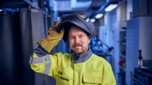 Työntekijä suojavarusteissa, keltaisessa takissa ja hitsauskypärässä.