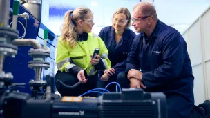 Työtakkinen nainen mittaustilanteessa, kaksi oppilasta katsovat vieressä