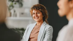 Hymyilevä nainen istuu vaaleassa takissa, tausta epätarkka.