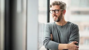 Mies katsoo ikkunasta ulos, harmaassa paidassa, rennon näköisenä, lasit päässä.