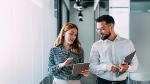 Kaksi ihmistä keskustelee käytävällä, pitelevät tabletteja.