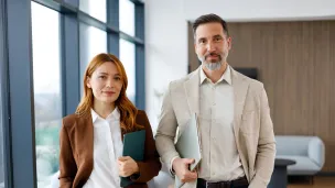 Kaksi henkilöä seisoo, pukeutuneet muodollisesti, kädessään kansiot, toimistoympäristössä.