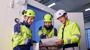 Kolme työntekijää huomiovaatteissa keskustelevat asiakirjasta sisätiloissa.