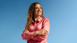 Smiling woman in pink shirt, arms crossed, against clear blue sky.