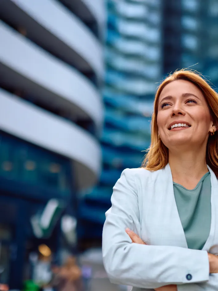 Hymyilevä nainen bleiserissä seisoo itsevarmasti kaupunkimaisemassa.
