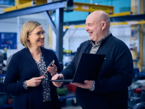 Kaksi hymyilevää henkilöä keskustelemassa teollisuusympäristössä, toinen pitää kannettavaa.