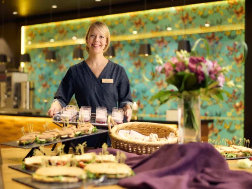 Hymyilevä nainen seisoo buffet-pöydän ääressä ja kukkia, voileipiä näkyy.