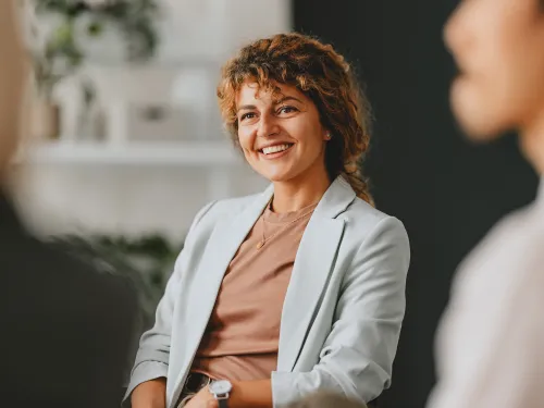 Hymyilevä nainen istuu vaaleassa takissa, tausta epätarkka.