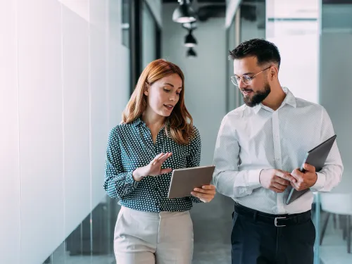 Two colleagues discuss while walking, holding a tablet and a folder.
